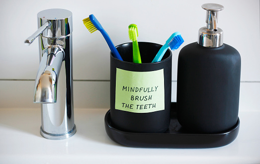 Close-up of a water tap, black soap dispenser and a cup for toothbrushes with a sticker containing a handwritten inscription: Mindfully brush the teeth. Mindfulness training concept
