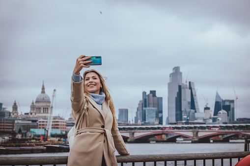 Candid portrait of a young cheerful blond girl in her 30s taking selfie with her mobile phone while smiling, the background is city of London's skyline, England, UK. Selective focus on the model with plenty of copy space at the background and sky, which is defocused Saint Paul’s cathedral, Blackfriars Bridge and all modern business buildings at the heart of the capital. Photo created during cold season outdoors and the model is with warm casual clothes on a cloudy day  - creative stock photo