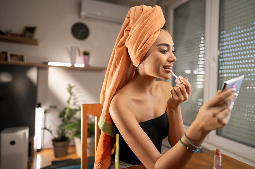 Beautiful young woman wearing towel wrapped around hair sitting at the table and applying lipstick in the morning