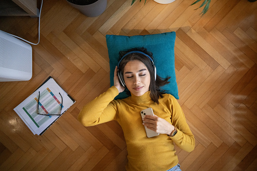 High angle view of relaxed young woman lying on floor and listening music on wireless headphones while taking break from learning at home