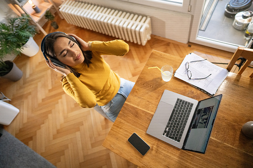 High angle view of beautiful young woman listening music on headphones while taking a break from work in home office