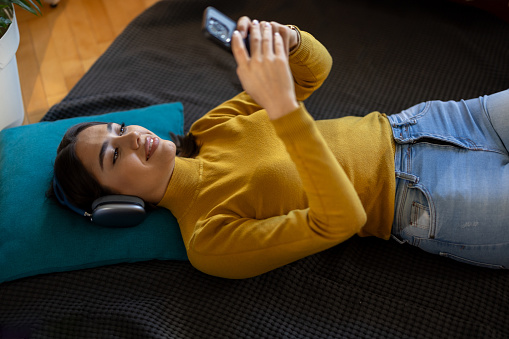 Smiling young woman relaxing while lying on floor and listening music on wireless headphones at home
