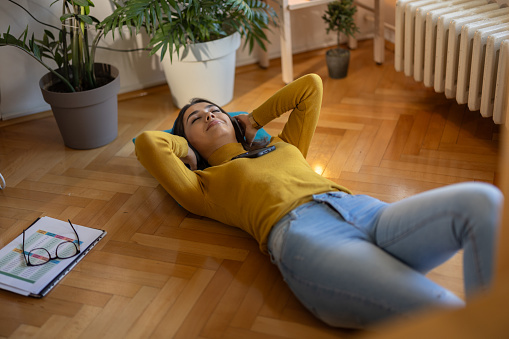 Young woman relaxing while lying on floor and listening music on wireless headphones while taking break from learning at home
