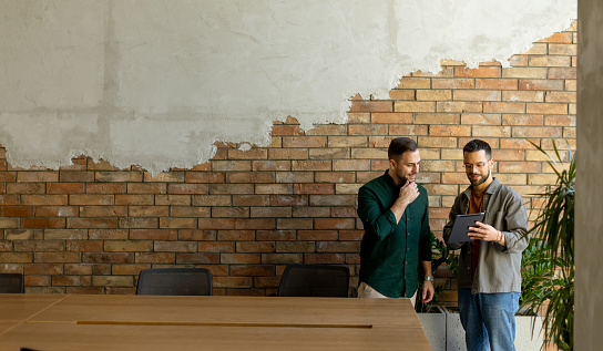 Two professionals are engaged in a focused conversation while holding digital tablet in a contemporary office space, featuring rustic exposed brickwork and warm natural light