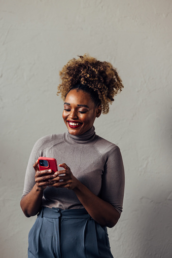 A smiling African-American entrepreneur using her smartphone while standing against grey background. (studio shot, copy space)