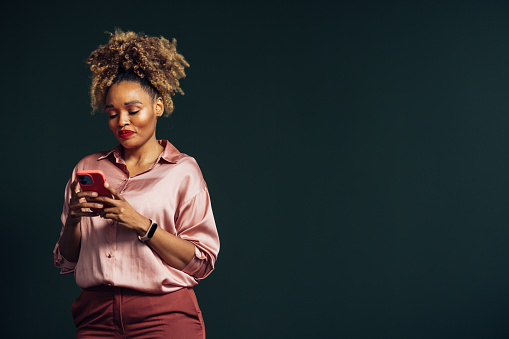 A smiling African-American entrepreneur texting on her smartphone while standing against grey background. (studio shot, copy space)