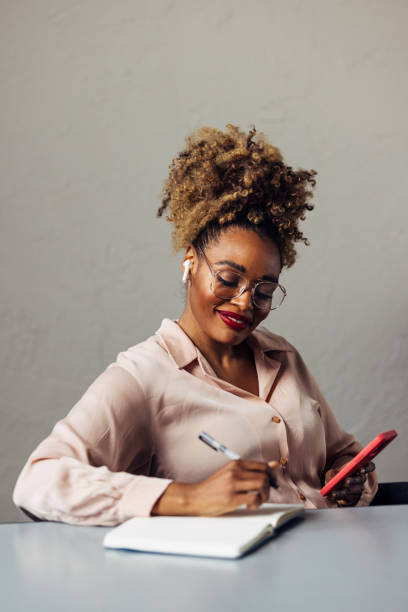 istock A Happy Beautiful Businesswoman With Blonde Curly Hair Taking Some Notes While Using Her Earbuds And Mobile Phone 2002295632