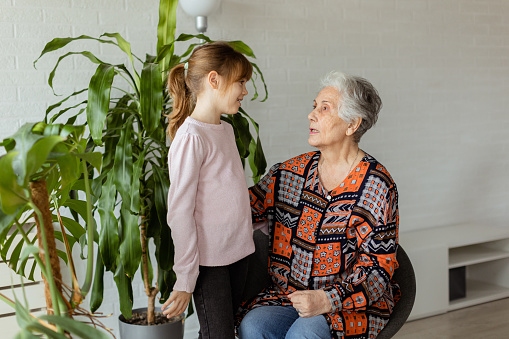 In a softly-lit room, a young girl stands beside a potted plant, sharing a moment of connection and conversation with her elderly grandmother, the joy and warmth between them palpable