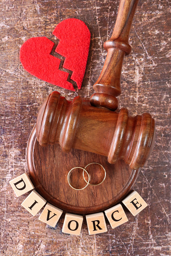 Stock photo showing close-up, elevated view of a row of letter dice spelling the word 'divorce' besides a hardwood ceremonial mallet and circular sound block with red, broken heart-shape and gold wedding bands. Divorce and relationship difficulties concept.