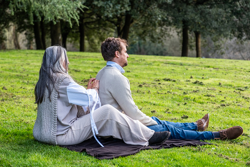 A man and a woman practising concentration exercises and spiritual connection sitting in a green meadow surrounded by trees in a park.