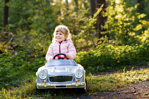 Little preschool girl driving big vintage toy car. Happy child having fun with playing outdoors. Active preschooler child enjoying warm autumn day in forest. Smiling stunning kid playing.