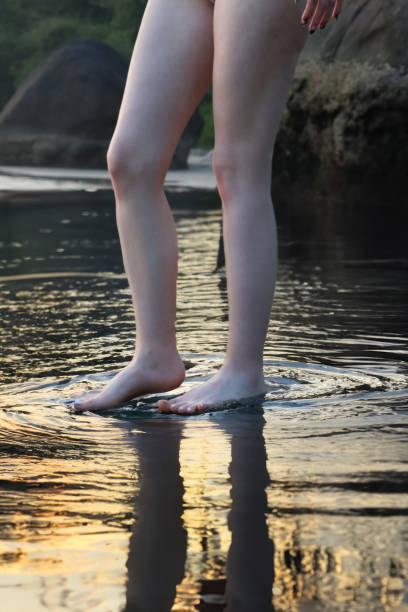 nahaufnahme einer nicht erkennbaren kaukasischen frau, die im meerwasser paddelt, gezeitenwellen, beine und nackte füße, die sich im seichten wasser spiegeln, sandstrand am meer bei sonnenuntergang, fokus auf den vordergrund - human foot wading sea human toe stock-fotos und bilder