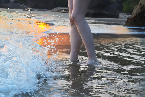 Stock photo showing an unrecognisable, caucasian woman paddling in the gentle sea waves, showing her barefoot partly in the water as tide comes in and the sea splashes over them.