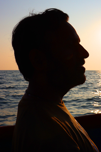 Stock photo showing close-up, profile view of unrecognisable male tourist, in silhouette, travelling on calm sea on a pleasure tour boat at sunset.