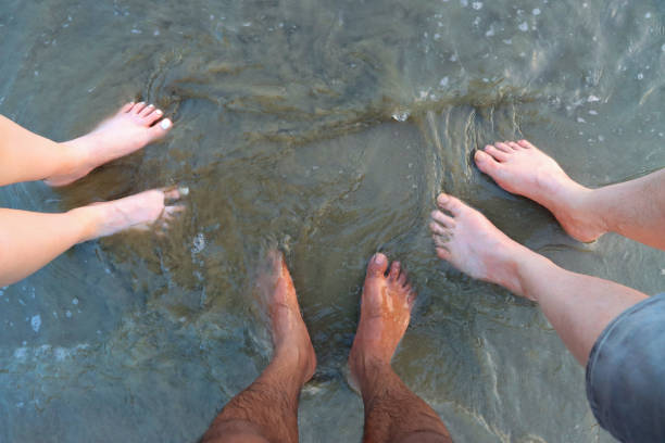Stock photo showing elevated view of three unrecognisable people standing barefoot on compact wet sand as tide rolls in. An Asian man stood in the middle of a caucasian man and a white woman. Racial equality concept