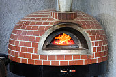 Close-up image of traditional brick wood fired pizza oven with stainless steel chimney flue, wood burning stove Italian restaurant pizzeria with burning logs and roaring orange flames, focus on foreground