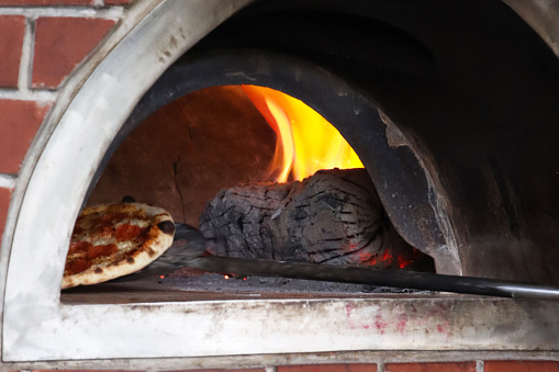 Stock photo showing close-up view of a traditional brick oven for cooking pizzas at an Italian pizzeria restaurant, brick-built wood burning stove with orange flames burning logs.  A long handled metal pizza paddle shovel can be seen removing a cooked pizza from the hot oven.
