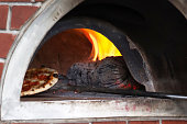 Full frame image of metal pizza paddle removing cooked pizza from traditional brick wood fired pizza oven with stainless steel chimney flue, wood burning stove Italian restaurant pizzeria with burning logs and roaring orange flames, focus on foreground
