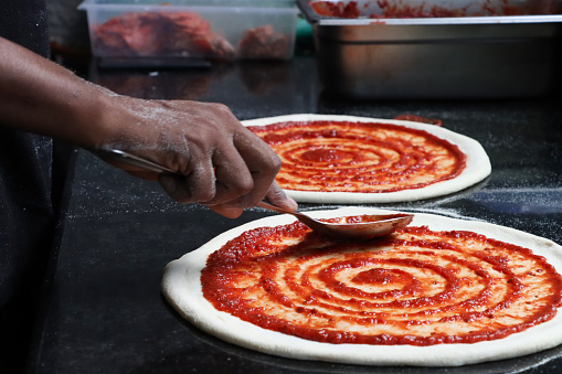 Stock photo showing close-up view of freshly prepared pizza dough base being covered with a rich tomato sauce with a metal serving spoon, by the hand of an unrecognisable pizza chef (Pizzaiolo), before having grated mozzarella cheese and torn basil leaves added to make a Margherita pizza.