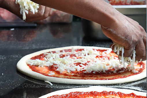 Stock photo showing close-up view of freshly prepared pizza dough base covered with rich tomato sauce having grated mozzarella cheese added by hand of unrecognisable pizza chef (Pizzaiolo).