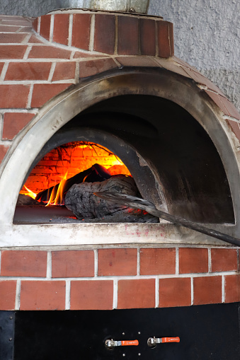 Stock photo showing close-up view of a traditional brick oven for cooking pizzas at an Italian pizzeria restaurant, brick-built wood burning stove with orange flames burning logs.  A long handled metal pizza paddle shovel can be seen being removed after placing a pizza into the hot oven.