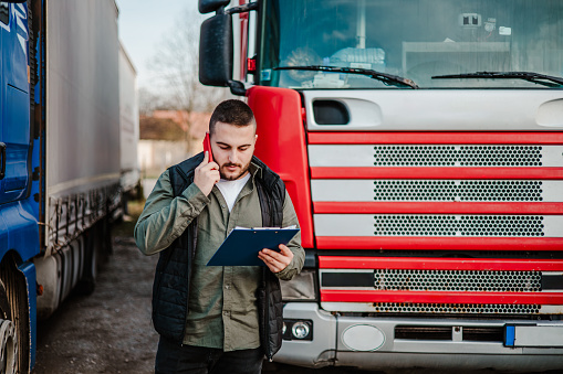 Young handsome truck driver standing in front of his truck and using smart phone for communication.