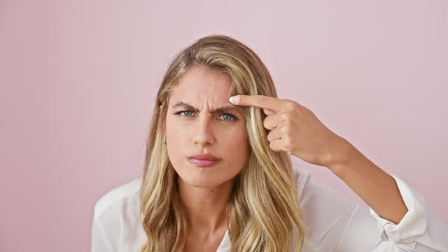 Unhappy young blonde woman in shirt, pointing at ugly acne blackhead on forehead, over isolated pink background - skin problem nightmare