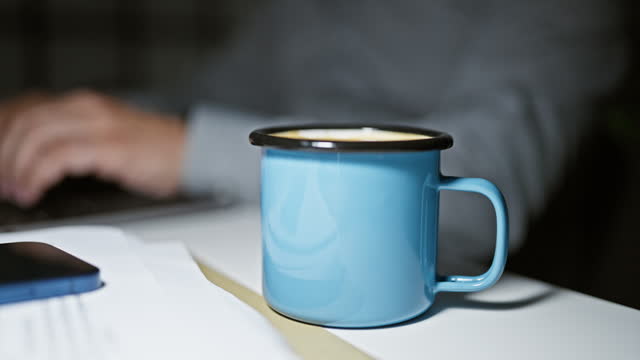 Close-up of a blue coffee mug on a white table with out-of-focus man typing on a laptop in the dark office background.