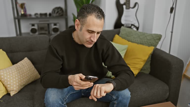 A focused man checks the time on his smartwatch while holding a phone in a cozy living room.