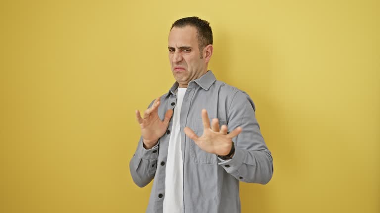 Young hispanic man standing, wearing shirt over yellow isolated background, face displaying disgusted expression. fearful reaction, hands raised due to aversion and displeasure.