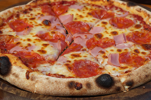 Stock photo showing close-up, elevated view of wooden pizza paddle displaying a sliced ham and pepperoni pizza, topped with a rich tomato sauce and melted buffalo mozzarella.