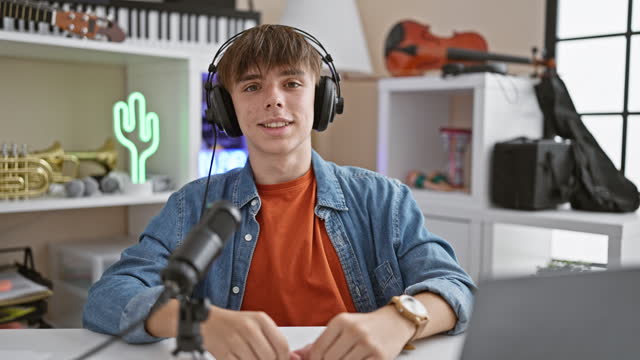 A young caucasian male smiles engagingly while recording in a home studio setup with a neon cactus and musical instruments in the background.