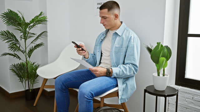 A young man in jeans and a denim shirt checks his phone and reads a document in a white room with modern chairs and green plants.