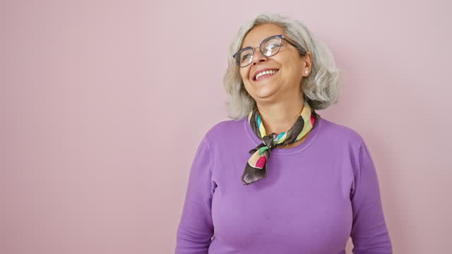 Confident grey-haired woman with glasses stands laughing, a beaming smile on her face as she looks off to the side over a pink isolated background, emulating great natural expression.