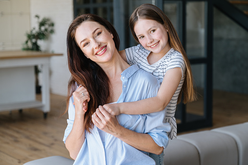 Happy woman and daughter spending time together, playing and resting in home on weekend day. Smiling girl child embracing mother and looking at camera. Playful mood concept