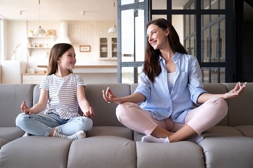 Mental health, calmness and mindfulness concept. Joyful woman teaching meditation kid, sitting in lotus pose with little daughter on sofa in living room. Child repeating yoga exercises after mother
