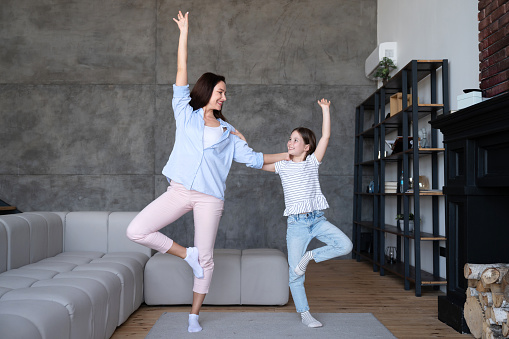 Smiling mother and daughter in casual wear standing in cozy living room at home and practicing yoga. Spending time together on weekend. Lifestyle concept