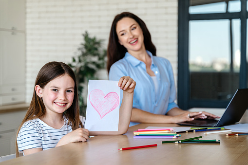 Happy smiling child holding postcard with red heart drawing with pencils. Kid sitting at table in dining room at home looking at camera, while mother working at notebook.