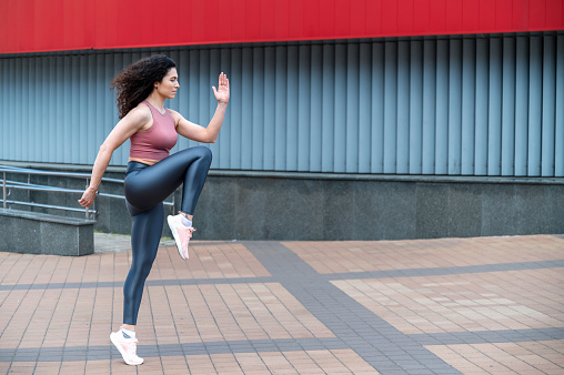 Warm-up, doing cardio workout outdoor. Sporty young woman in sportswear standing on one leg and touching knee with elbow, doing high knees exercise on street.