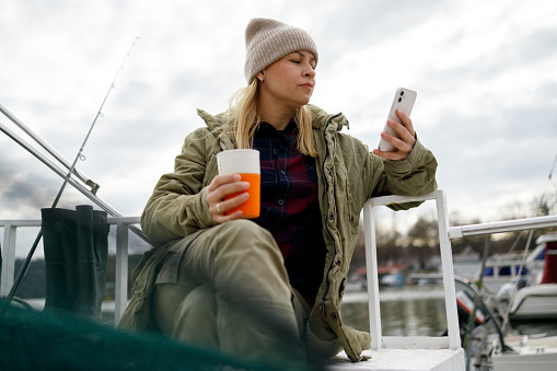 A fisherwoman sits on the deck of her boat, holding a reusable mug with an orange band in one hand and checking her smartphone with the other, possibly reviewing weather conditions or communicating with fellow anglers, with fishing gear and the marina in the background.