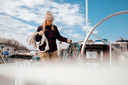 A European woman in a warm beanie and plaid shirt ties a rope on a sunny day at the marina during the winter season.