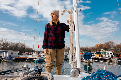 A European woman in a warm beanie and plaid shirt ties a rope on a sunny day at the marina during the winter season.