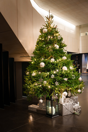 Elegant Christmas tree with white and silver ornaments, warm lights, and a star topper in a modern interior, surrounded by stylishly wrapped gifts and a candle lantern.