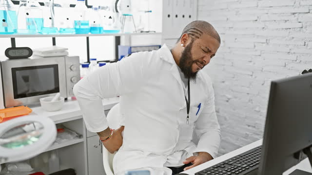 Man in laboratory wincing in pain while holding his lower back, indicating a workplace injury or discomfort.