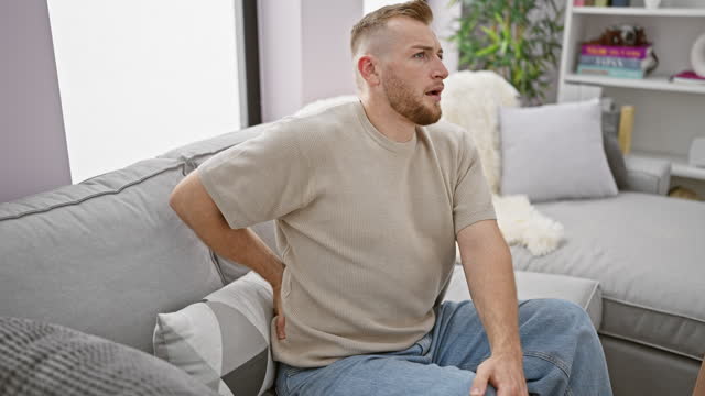 Attractive young man with beard in pain sitting on a gray couch at home holding lower back.