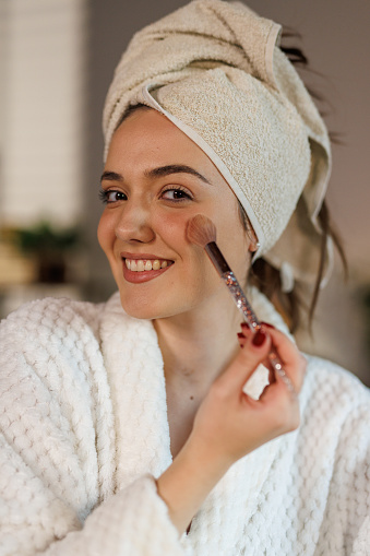 Portrait of beautiful young woman sitting at dining table, in a bathrobe and head wrapped in a towel, fresh out of shower, applying face make up when getting ready for a night out. She is looking at camera as if it is a mirror. Point of view.