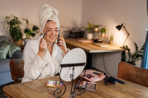 Portrait of beautiful young woman sitting at dining table, in a bathrobe and head wrapped in a towel, fresh out of shower, applying face make up when getting ready for a night out. She is also talking on the phone with a friend.