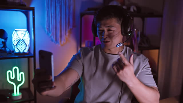 A young asian man takes a selfie in a neon-lit gaming room at night, highlighting technology and lifestyle.