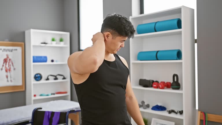 A young asian man grimaces in pain, holding his neck while standing in a modern rehabilitation clinic.