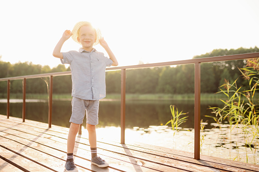 A happy boy in a hat stands on the embankment near the lake at sunset, enjoys the view of the calm lake and forest, breathes fresh clean air. Vacation, summer concept.